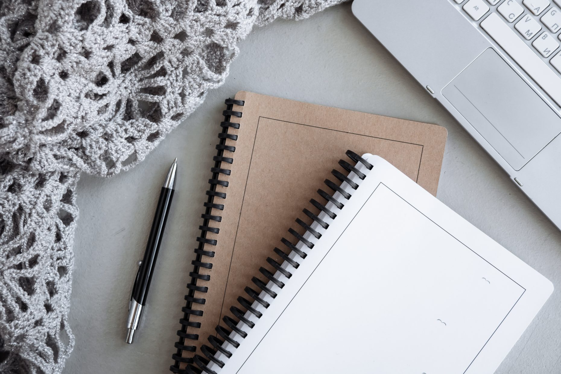 Top-down view of kraft and white spiral notebooks, a pen, a silver laptop corner and grey knit fabric