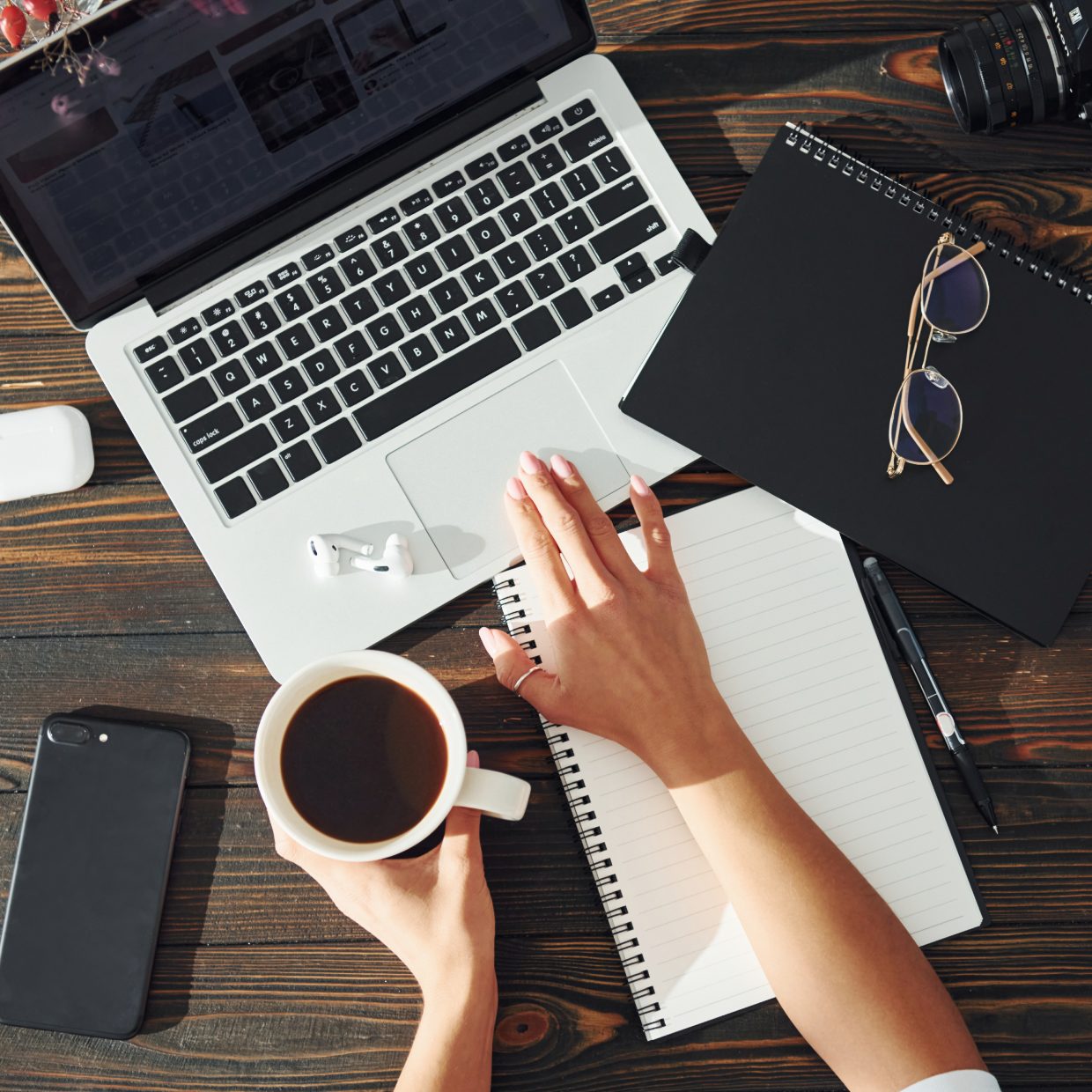 Top-down view of a laptop, black coffee, spiral notebook, phone and glasses on a dark wooden desk